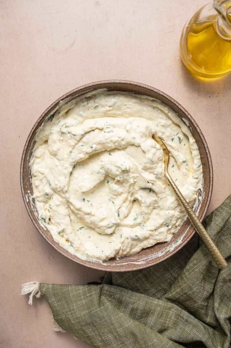 A bowl of creamy, herbed ricotta filling with a spoon, set on a light surface next to a green napkin and a bottle of olive oil.