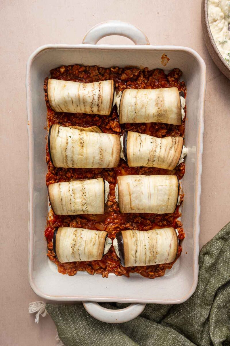A baking dish with eight rolled eggplant slices filled with cheese placed on top of a layer of tomato and ground lamb sauce, ready for baking.