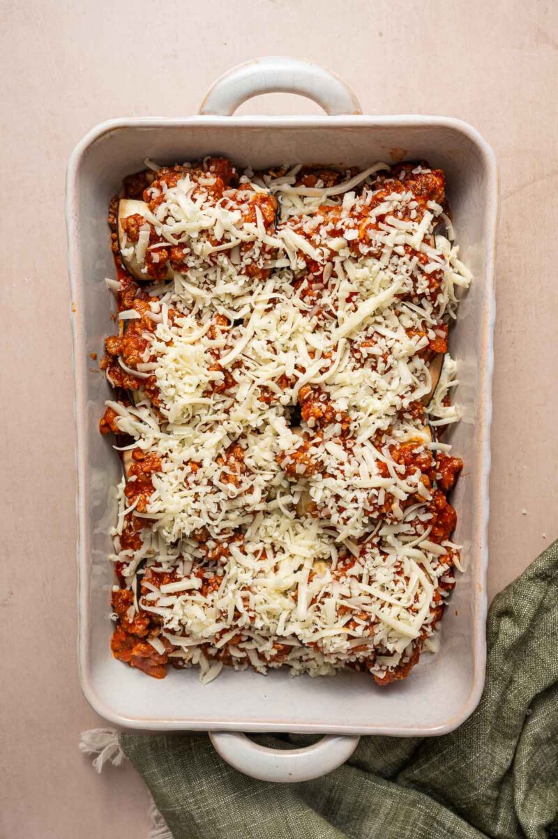 Rectangular un-baking dish filled with eggplant rollatini layered in tomato sauce and topped with shredded cheese, placed on a light surface next to a green cloth.