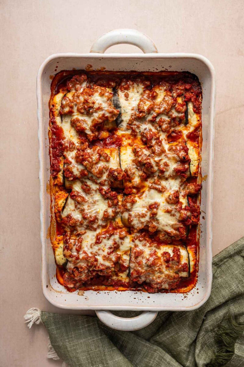 A baked casserole dish filled with rolled eggplant covered in ground lamb tomato sauce, ground meat, and melted cheese, set on a light surface with a green cloth beside it.