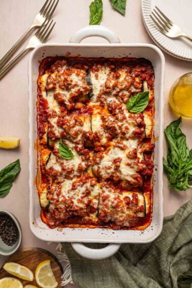 A baked casserole dish filled with rolled eggplant topped with ground lamb tomato sauce, melted cheese, and fresh basil, set on a table with plates, utensils, lemon wedges, and basil leaves.