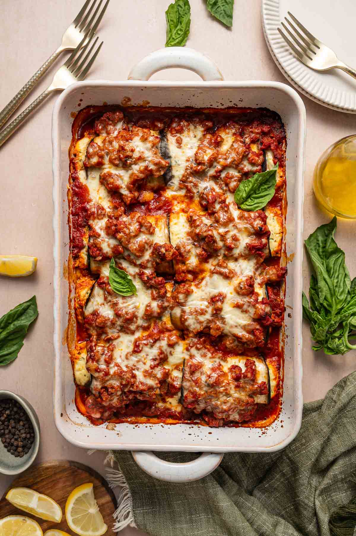 A baked casserole dish filled with rolled eggplant topped with ground lamb tomato sauce, melted cheese, and fresh basil, set on a table with plates, utensils, lemon wedges, and basil leaves.