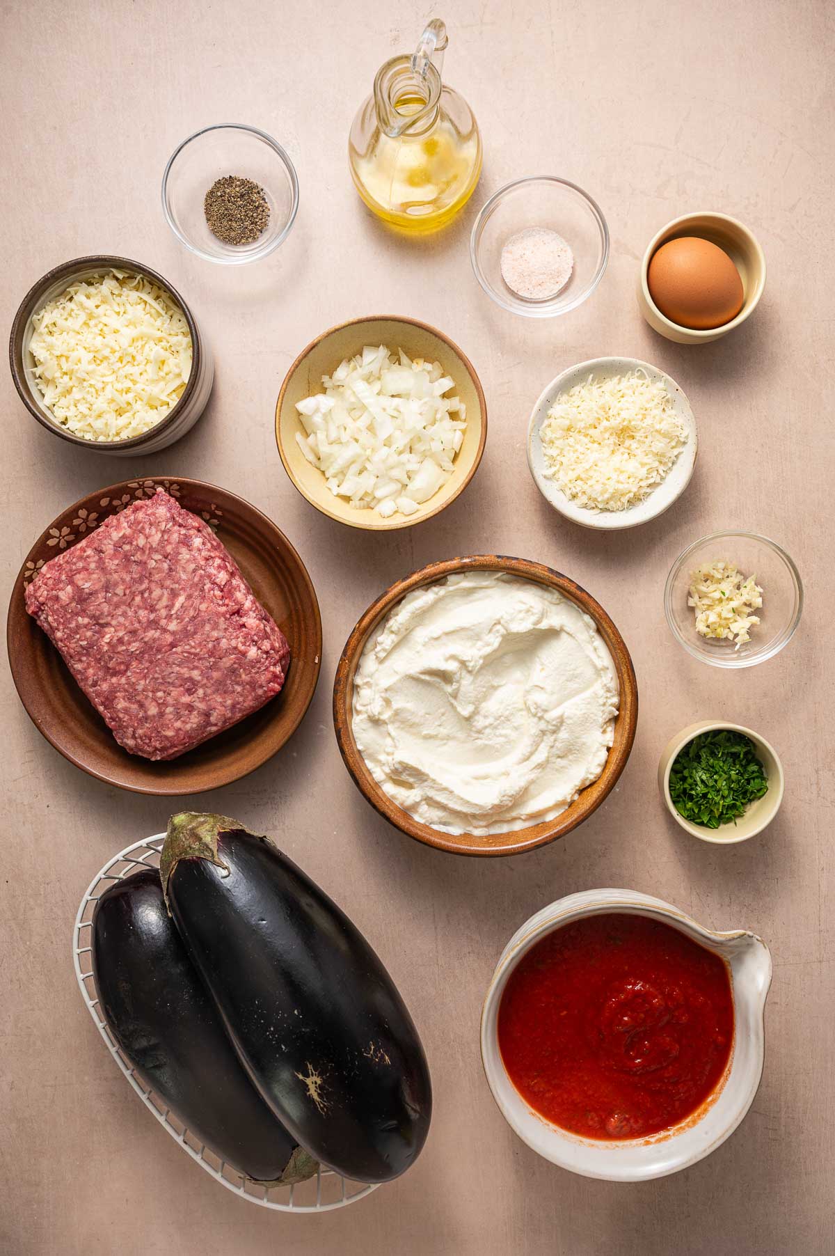 Ingredients for an eggplant rollatini arranged on a table, including ground lamb, eggplants, cheeses, ricotta, chopped onion, sauce, egg, oil, herbs, and seasonings in bowls.
