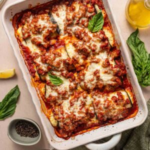 A baked dish of cheesy, tomato-sauce-covered stuffed eggplant rollatini in a white casserole dish, garnished with fresh basil leaves. Lemon wedges and pepper are nearby.