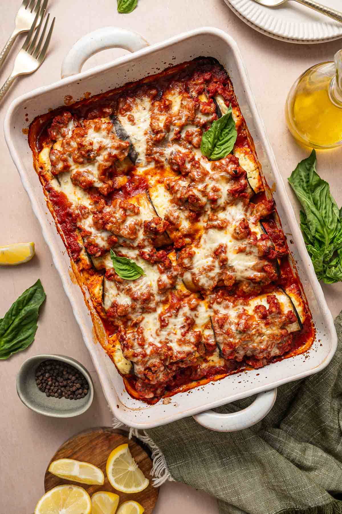 A baked dish of cheesy, tomato-sauce-covered stuffed eggplant rollatini in a white casserole dish, garnished with fresh basil leaves. Lemon wedges and pepper are nearby.