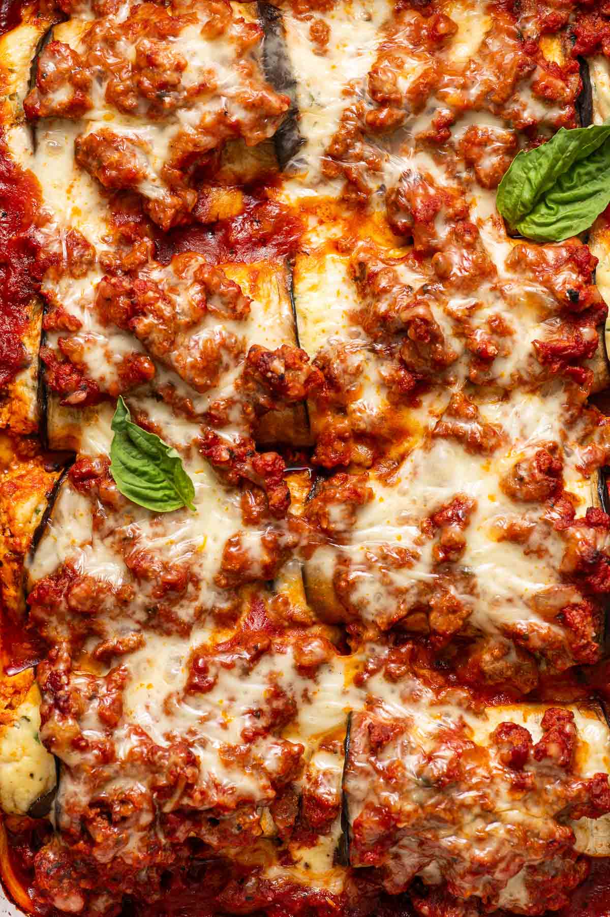 Close-up of baked eggplant rollatini topped with melted cheese, tomato sauce, ground meat, and garnished with fresh basil leaves.