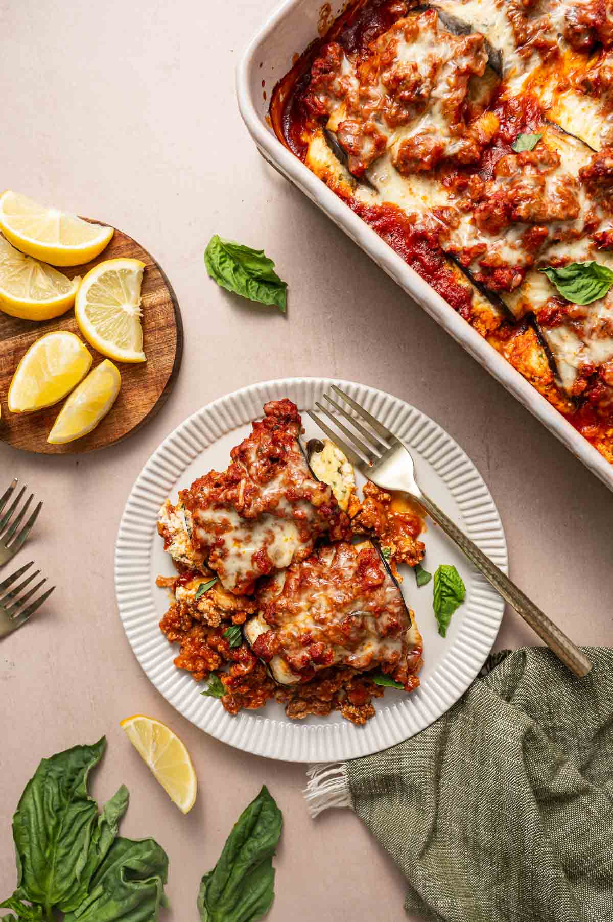 A plate of baked eggplant rollatini served with a fork, garnished with basil leaves and lemon wedges, next to a casserole dish of lasagna and a small plate with lemon slices.