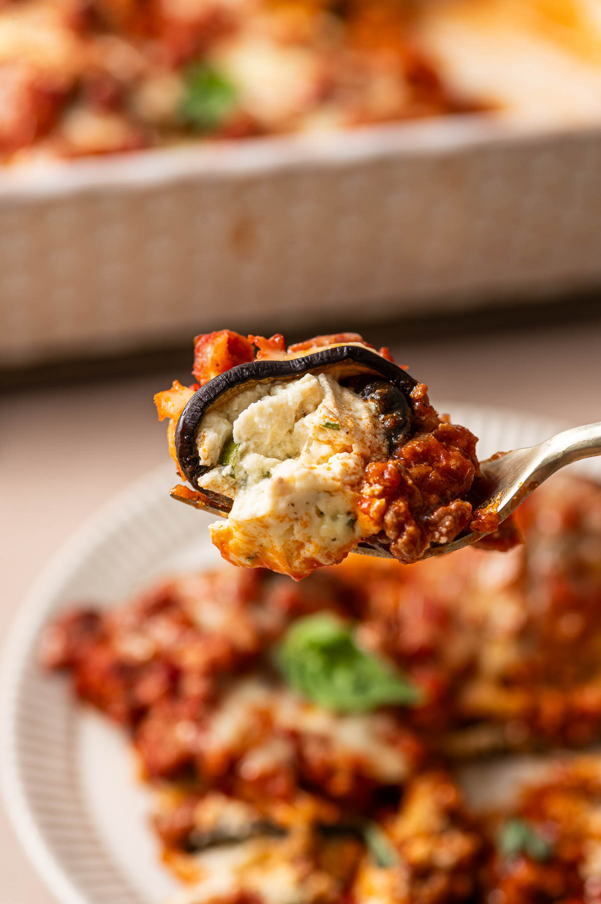 A fork holds a bite of baked eggplant rollatini layered with ricotta cheese and tomato sauce, with a plate of more in the background.