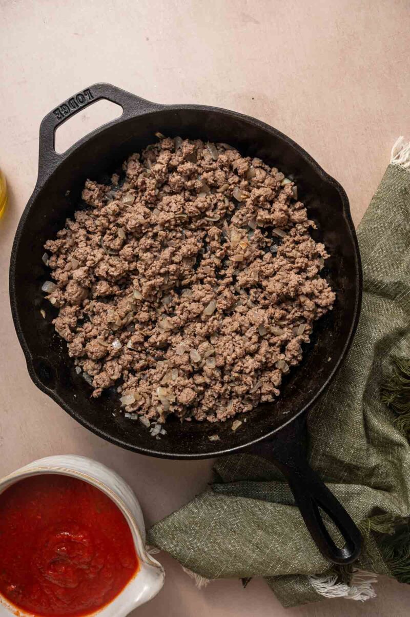 Cooked ground lamb with chopped onions in a black cast iron skillet, placed on a beige surface next to a bowl of tomato sauce and a green cloth.