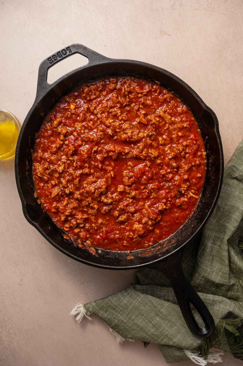 Cast iron skillet filled with ground lamb simmering in tomato sauce, resting on a light surface next to a green cloth and a small jar of olive oil.