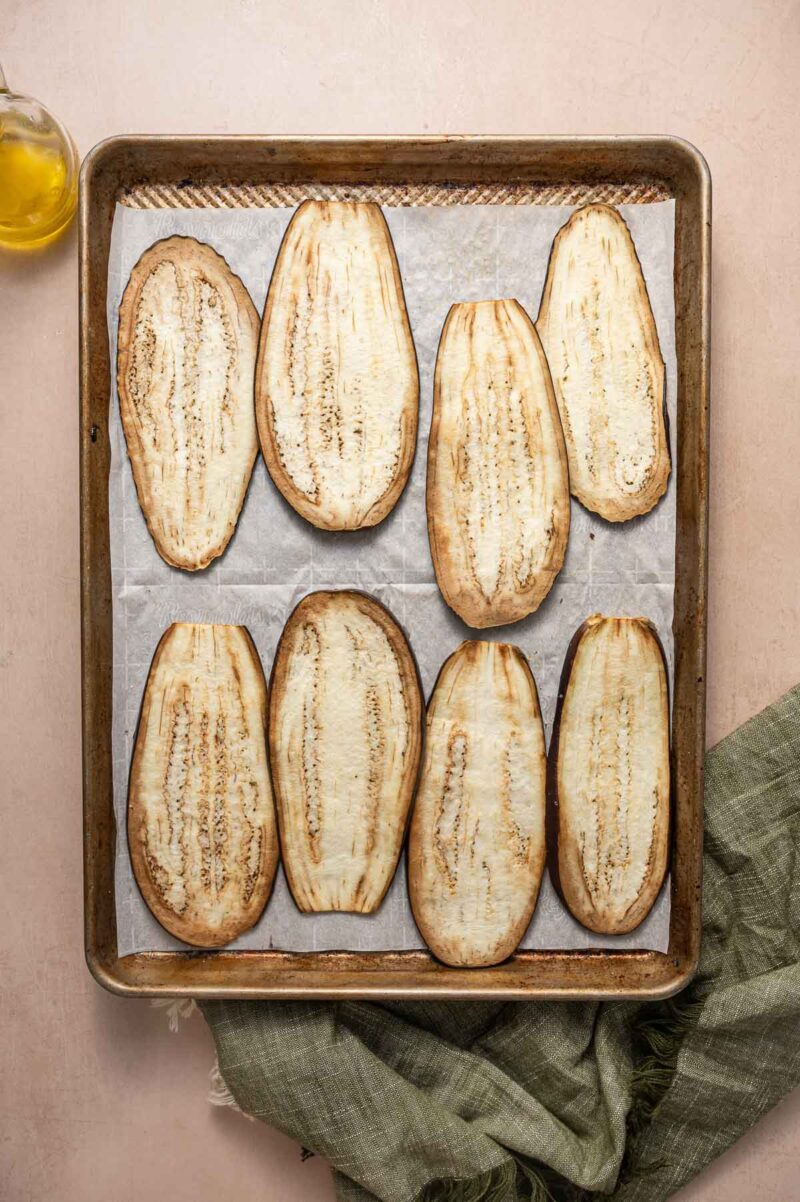 Eight slices of baked eggplant are arranged in a single layer on a parchment-lined baking sheet. A bottle of oil and a green cloth are nearby.