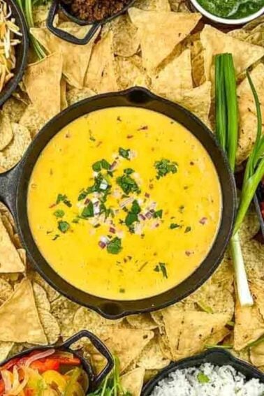 A platter with tortilla chips, cheese dip, shredded cheese, pico de gallo, shredded chicken, ground beef, rice, salsa, beans, sautรฉed peppers, and green onions.