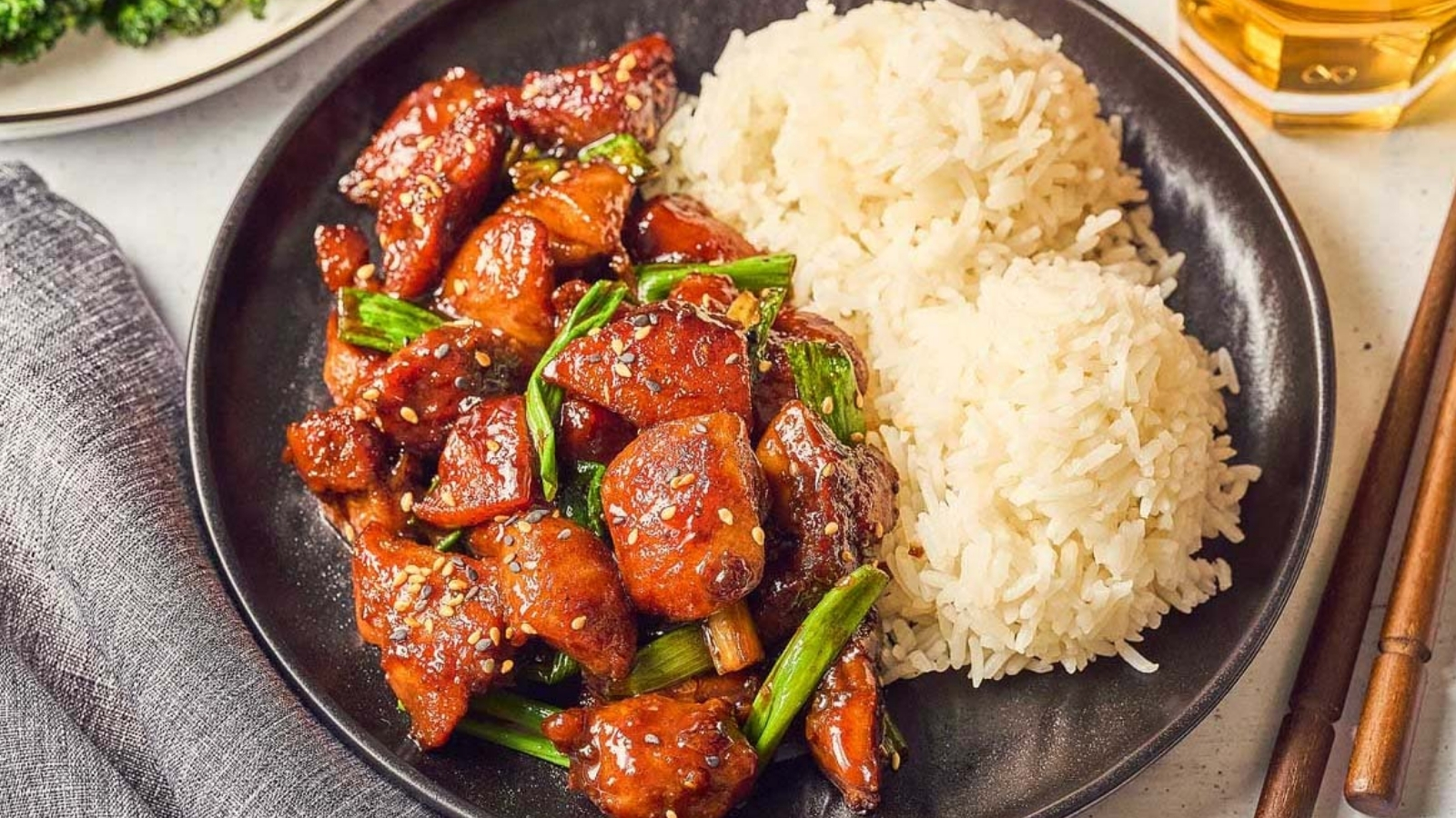 Black plate with glazed chicken pieces topped with sesame seeds and green onions beside two scoops of white rice, with chopsticks, a gray napkin, a glass with amber liquid, and a partially visible plate of broccoli nearby.