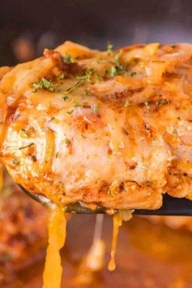 A close-up of a cooked, cheesy chicken breast with tomato sauce and herbs being lifted from a pan with a spatula.