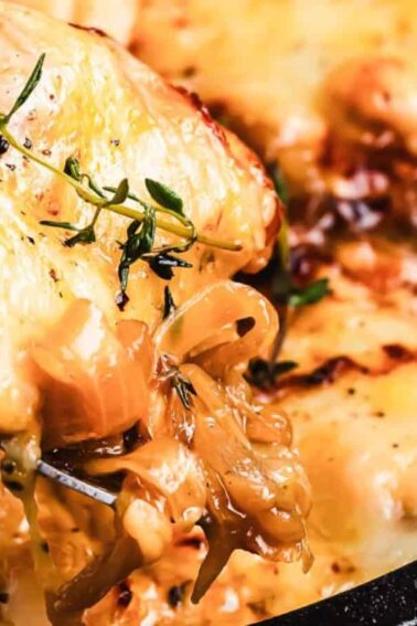 A close-up of a cheesy baked dish with caramelized onions, being served from a cast iron skillet with a metal spoon, garnished with fresh herbs.