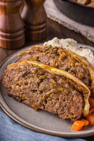 Two slices of French Onion meatloaf topped with caramelized onions, served with mashed potatoes and carrots on a gray plate.