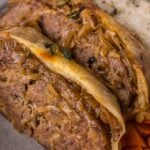 Two thick slices of French Onion meatloaf topped with caramelized onions, served with mashed potatoes and carrots on a plate.