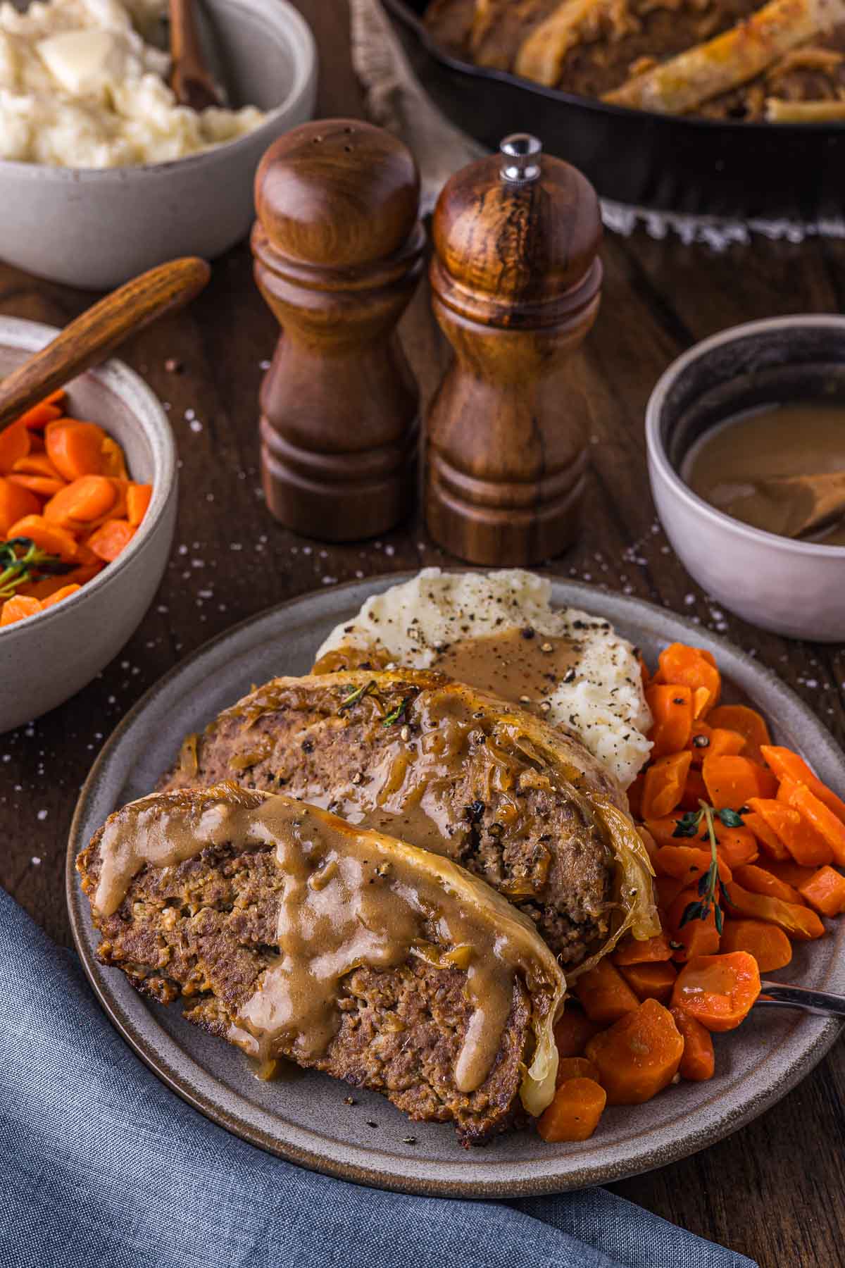 A plate with two slices of French Onion meatloaf topped with gravy, mashed potatoes with gravy, and cooked carrots. Salt and pepper shakers, gravy, and bowls of mashed potatoes and carrots are in the background.