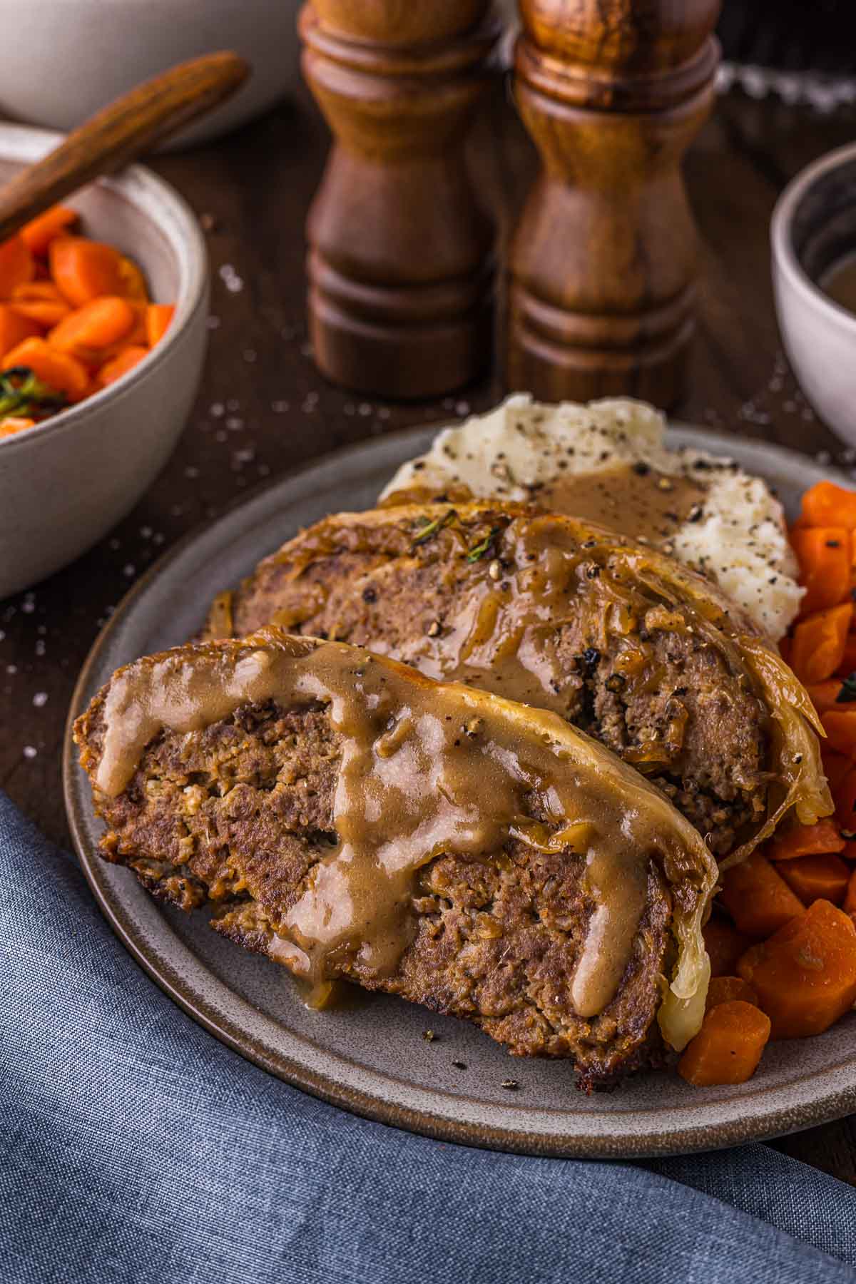 Two slices of French Onion meatloaf with brown gravy, served with mashed potatoes and cooked carrots on a plate. Salt and pepper shakers are in the background.