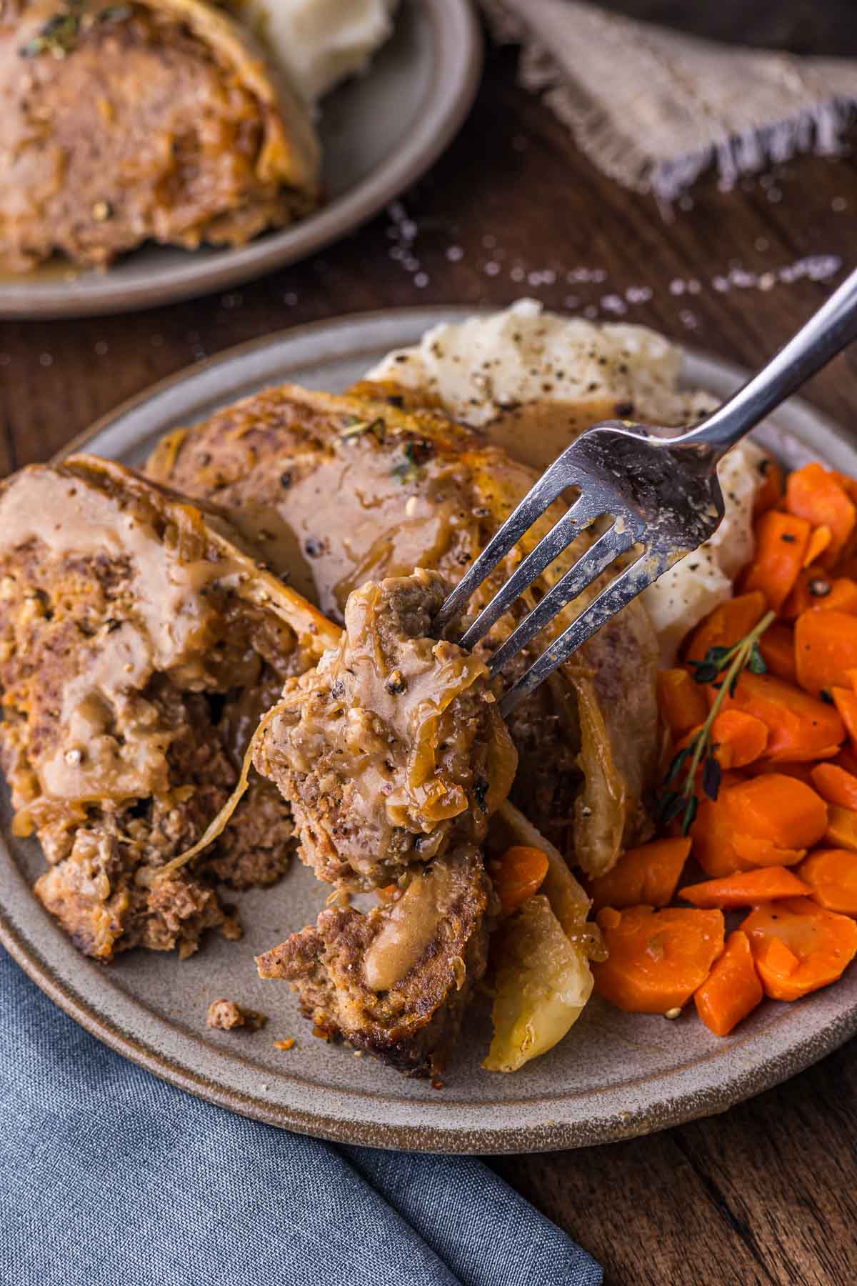 A plate with slices of French Onion meatloaf topped with gravy, mashed potatoes, and cooked carrots, with a fork holding a bite of meatloaf.