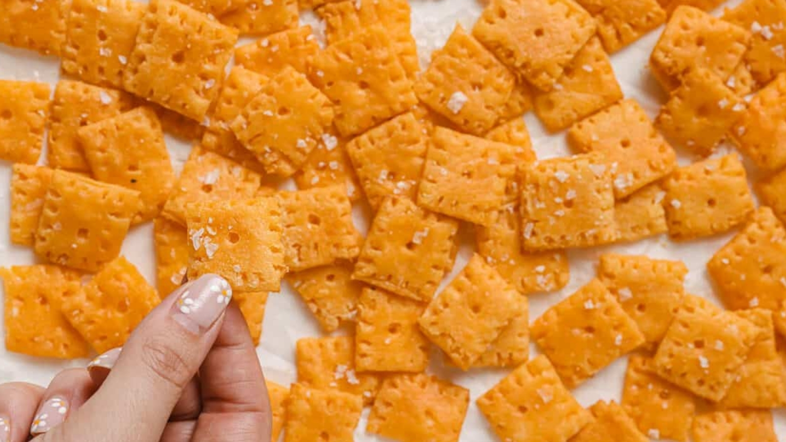 A hand holding a Cheez-It above a baking sheet of more crackers.