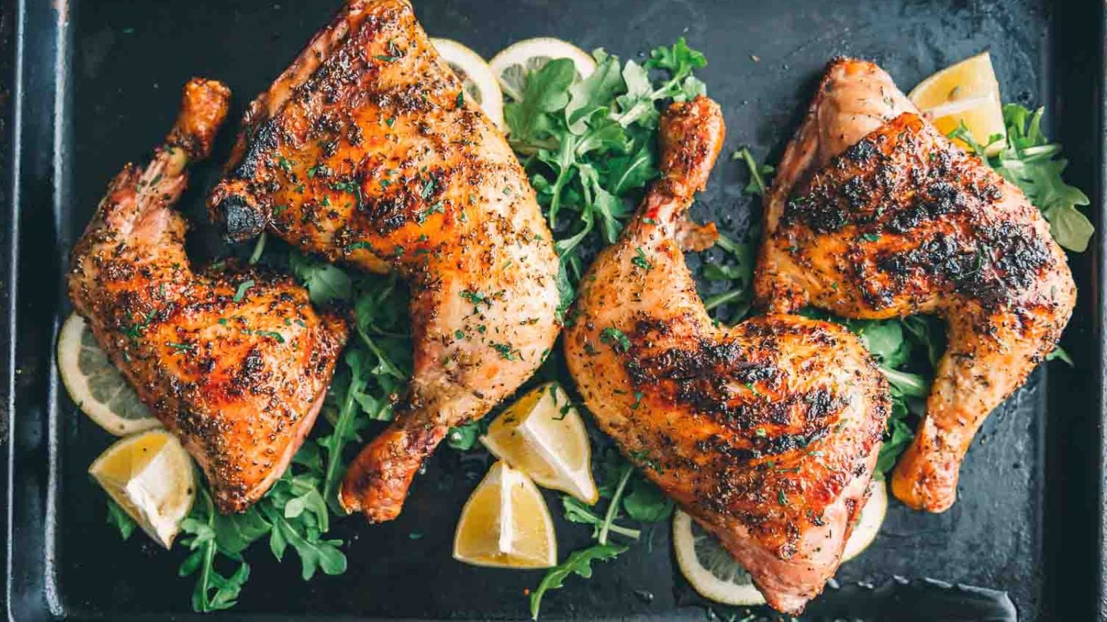 Four roasted chicken leg quarters garnished with chopped herbs, served on a bed of greens with lemon wedges on a black tray.