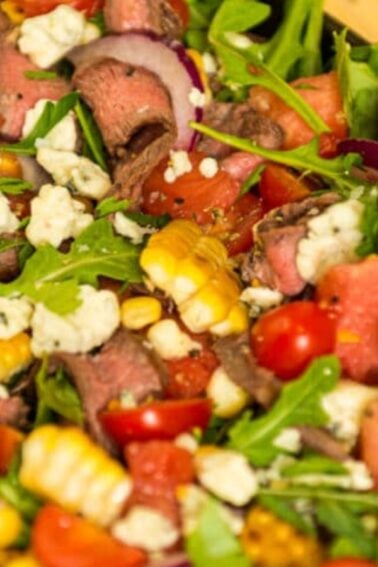 A vibrant salads platter with arugula, cherry tomatoes, corn, red onions, crumbled cheese, fresh watermelon slices, and roast beef artfully arranged on a wooden board.