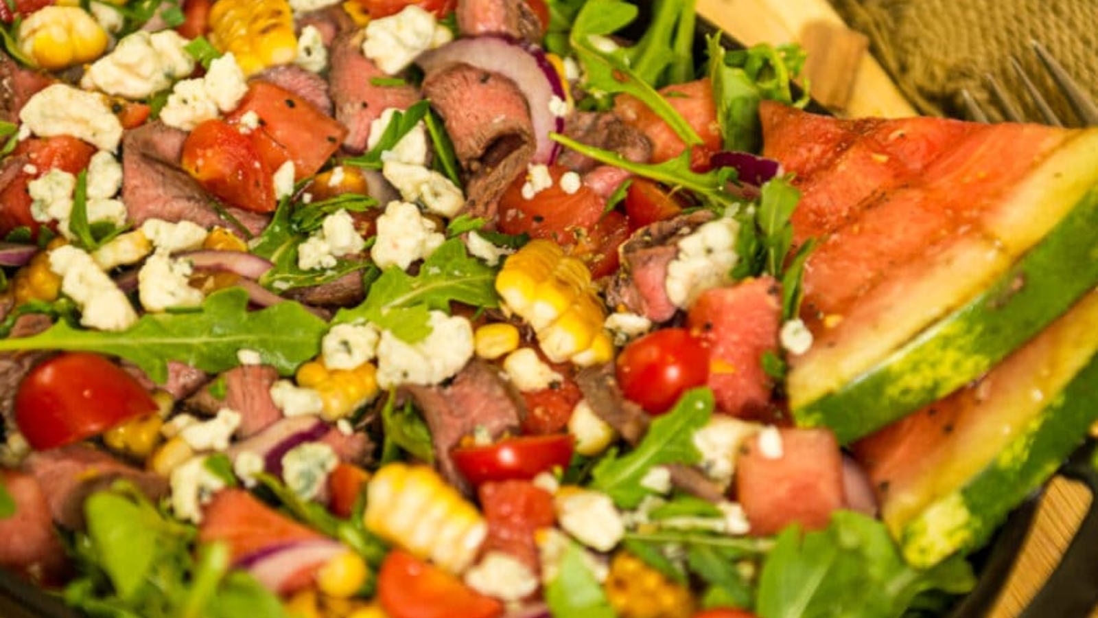 A vibrant salads platter with arugula, cherry tomatoes, corn, red onions, crumbled cheese, fresh watermelon slices, and roast beef artfully arranged on a wooden board.