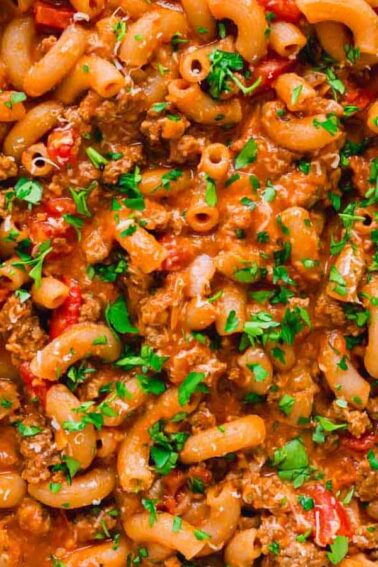 A skillet filled with macaroni pasta, ground meat, diced tomatoes, and sauce, garnished with chopped parsley.