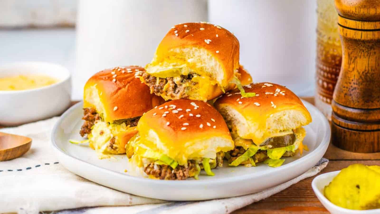 Four cheeseburger sliders with lettuce and pickles on a white plate, topped with sesame seed buns.