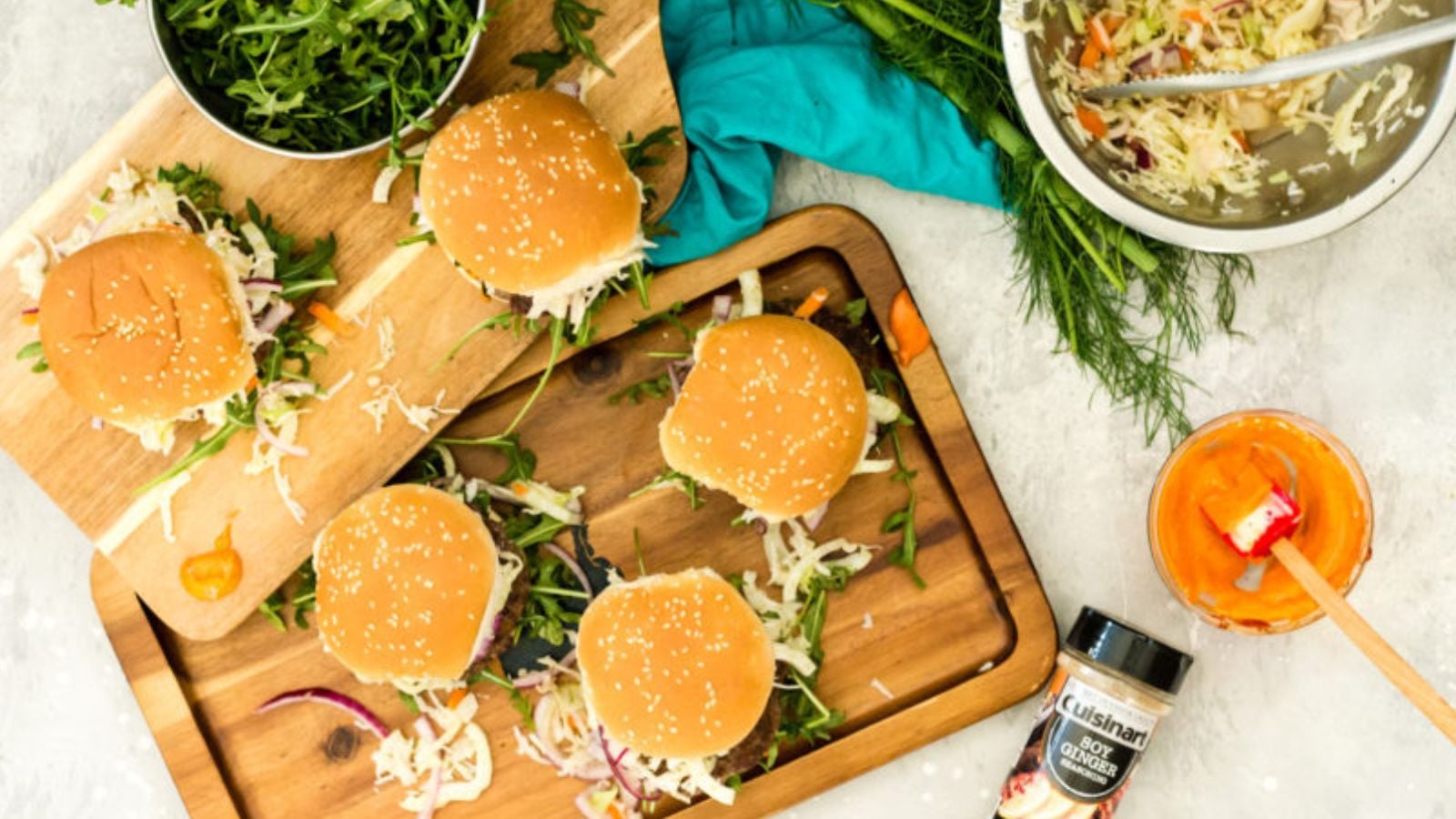 Five assembled burgers with sesame seed buns on wooden boards, surrounded by greens, a metal bowl of slaw, a jar of orange sauce, and a spice container on a light surface.