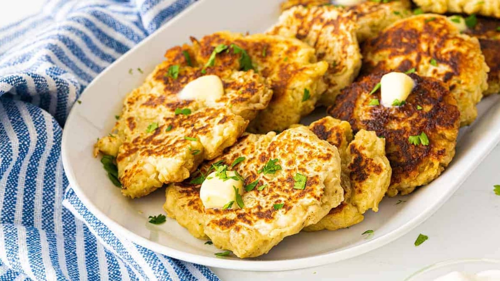 Oval plate filled with browned potato pancakes topped with small pieces of butter and chopped herbs, with a blue and white striped cloth beside the plate.