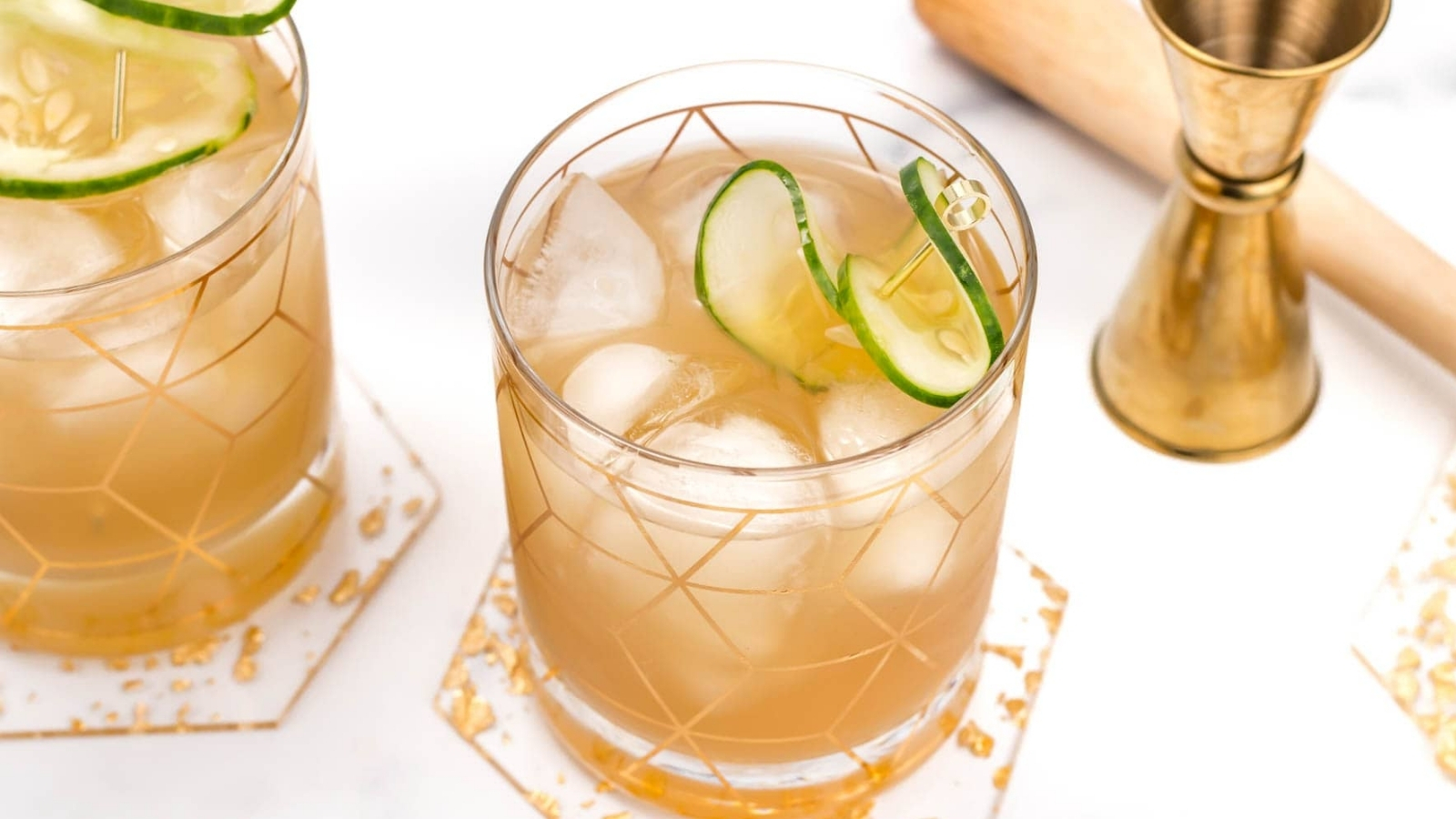 Two patterned glasses filled with a light brown drink and ice cubes are garnished with cucumber slices and set on hexagonal coasters, with a gold jigger and wooden utensil in the background.