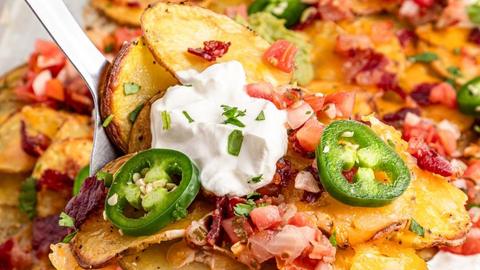 A metal spatula lifts a portion of sliced roasted potatoes topped with melted cheese, diced tomatoes, jalapeño slices, bacon pieces, sour cream, guacamole, and chopped herbs from a sheet pan.