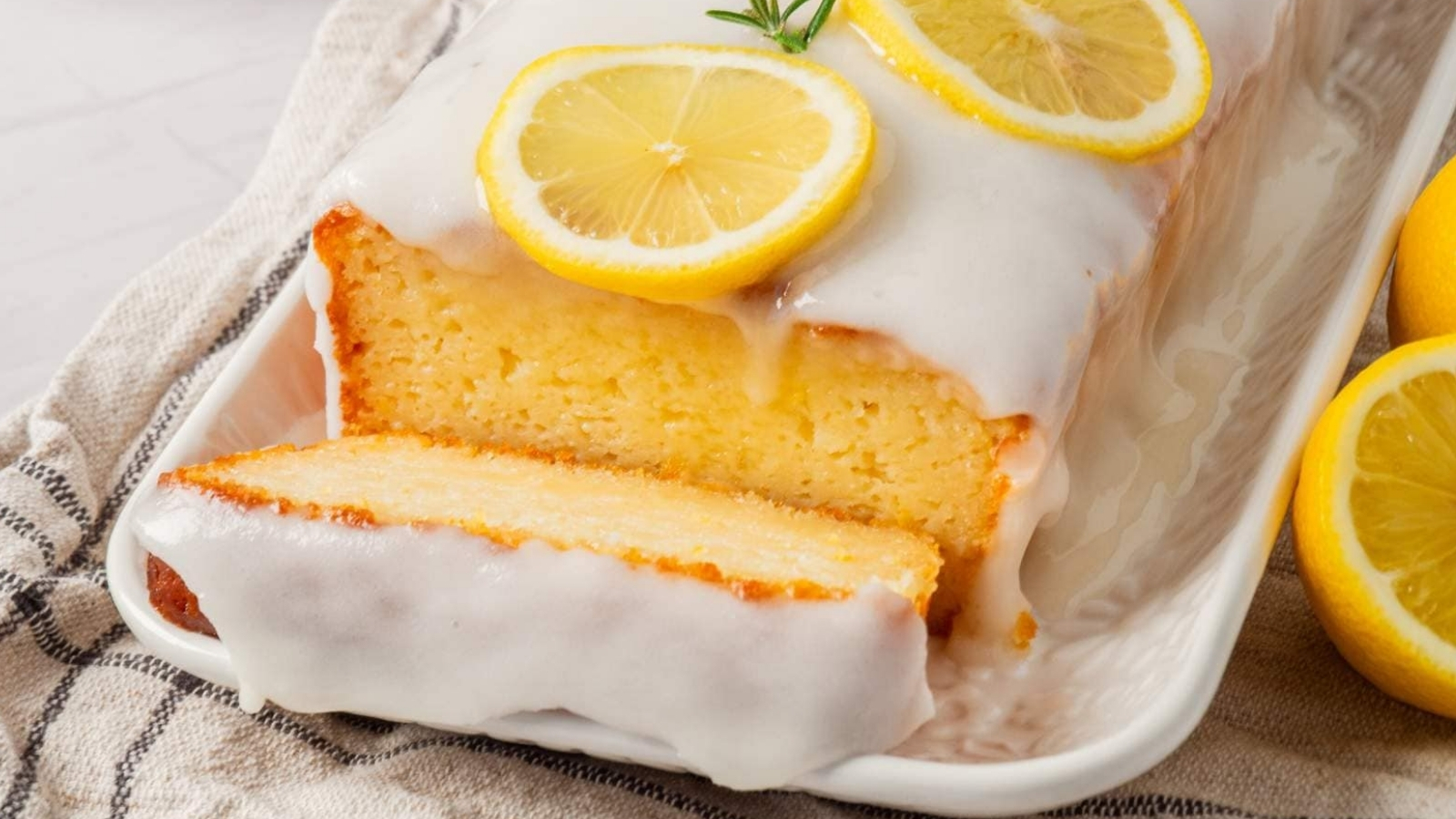 Lemon loaf cake on a white tray, topped with glaze and lemon slices, with a cut piece in the foreground and whole lemons nearby.
