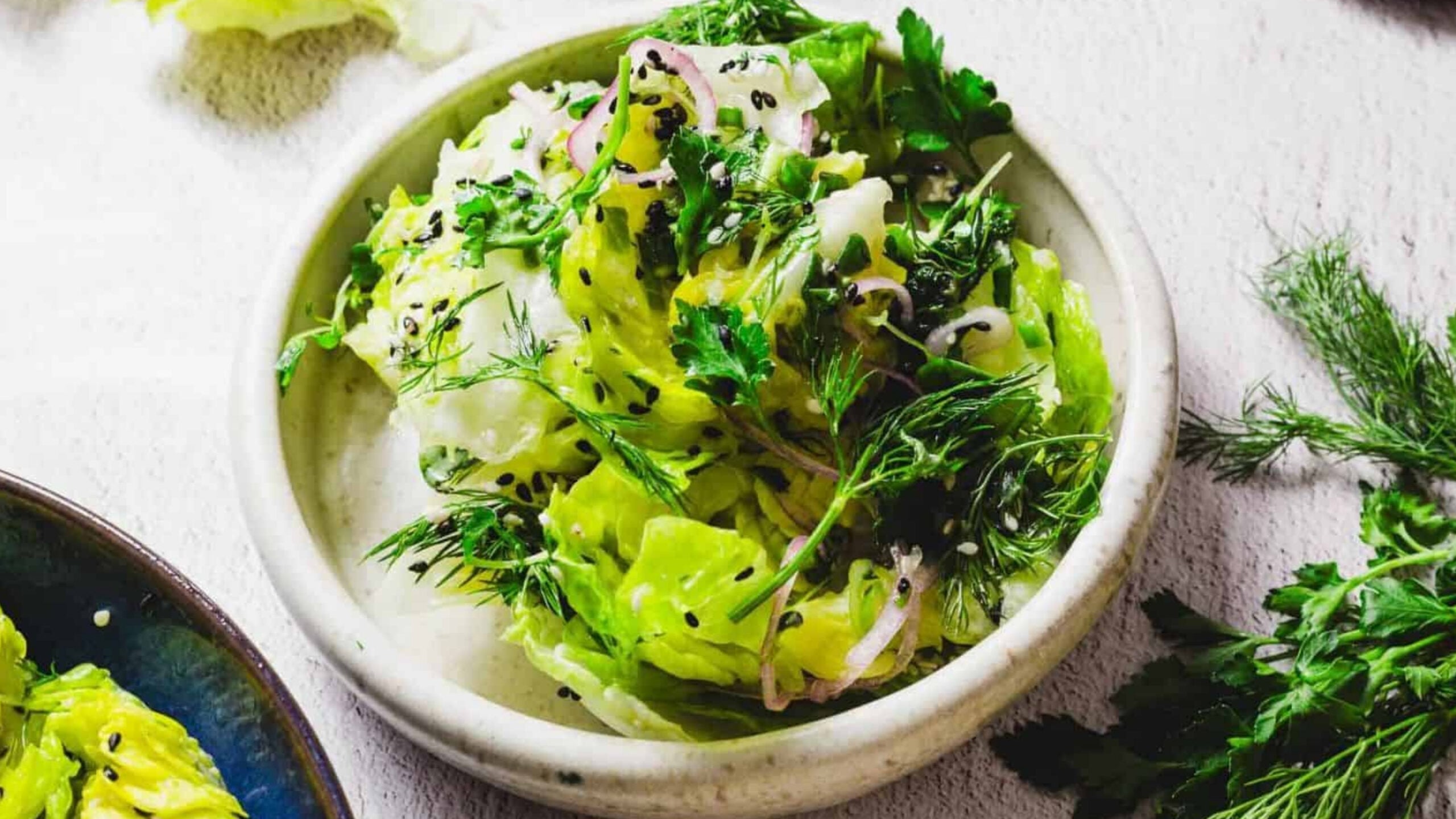 A bowl of salad containing leafy greens, fresh herbs, sliced onions, and black sesame seeds on a light surface. Another dish with similar contents is partially visible to the side. There are scattered herbs around the bowl.