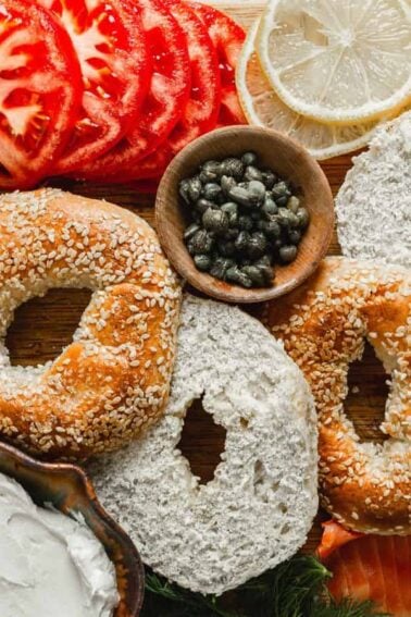 Assorted bagels with sesame seeds, cream cheese, smoked salmon, sliced tomatoes, cucumbers, red onions, lemon, dill, capers, and herbs arranged on a wooden board.