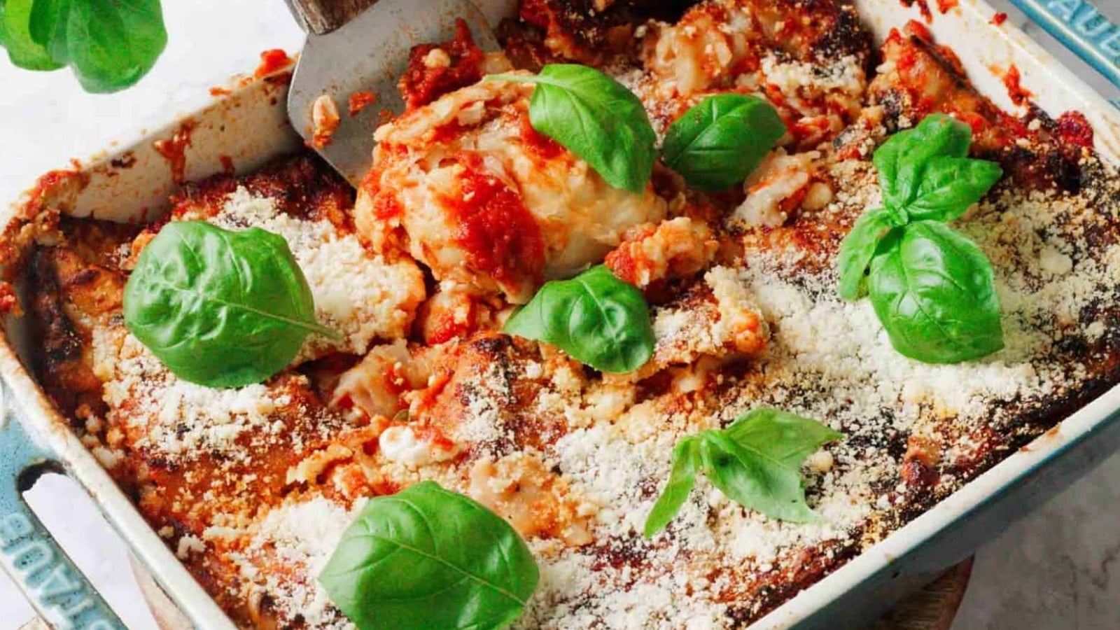 A baked lasagna in a rectangular dish, topped with tomato sauce, basil leaves, and grated cheese. A spatula is lifting a portion from the dish, revealing layers of pasta and filling. The dish is on a light countertop.