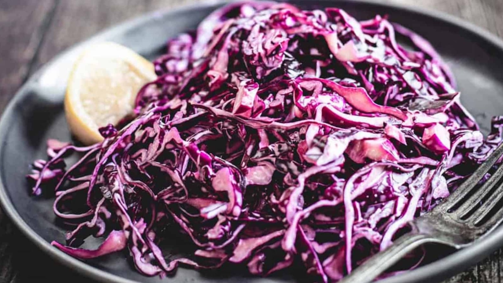 Shredded red cabbage salad on a dark plate with a lemon wedge and a fork.