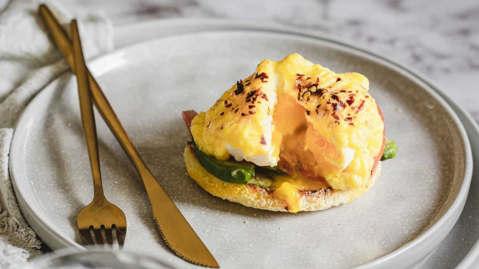 A plate with eggs benedict featuring a poached egg, hollandaise sauce, and avocado slices on an english muffin, garnished with spices, accompanied by gold cutlery. Perfect for Mother's Day recipes.