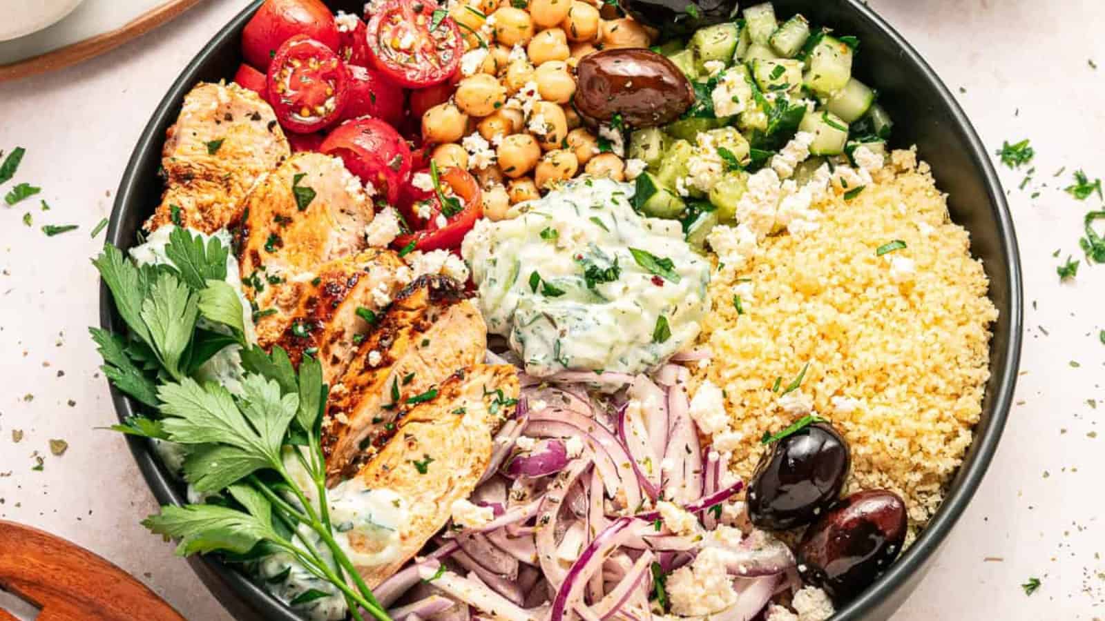 A colorful Mediterranean bowl with grilled chicken, couscous, tomatoes, cucumber, chickpeas, olives, and tzatziki.