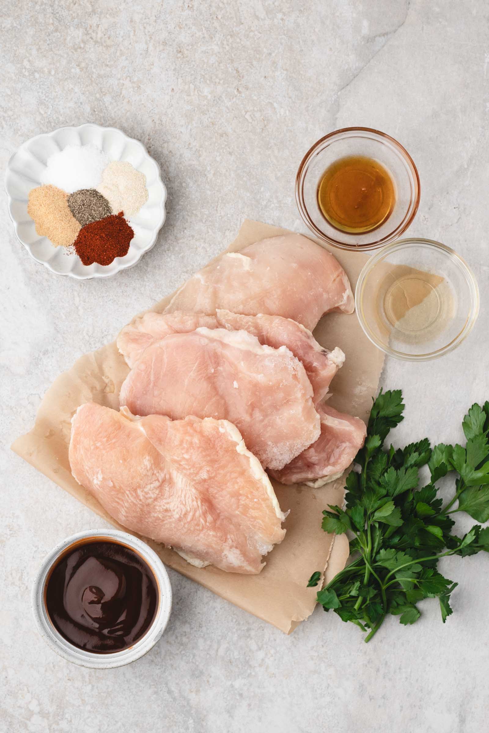 Raw chicken breasts on parchment paper surrounded by barbecue sauce, fresh parsley, two small bowls with liquids, and a dish of mixed spices on a light stone surface.