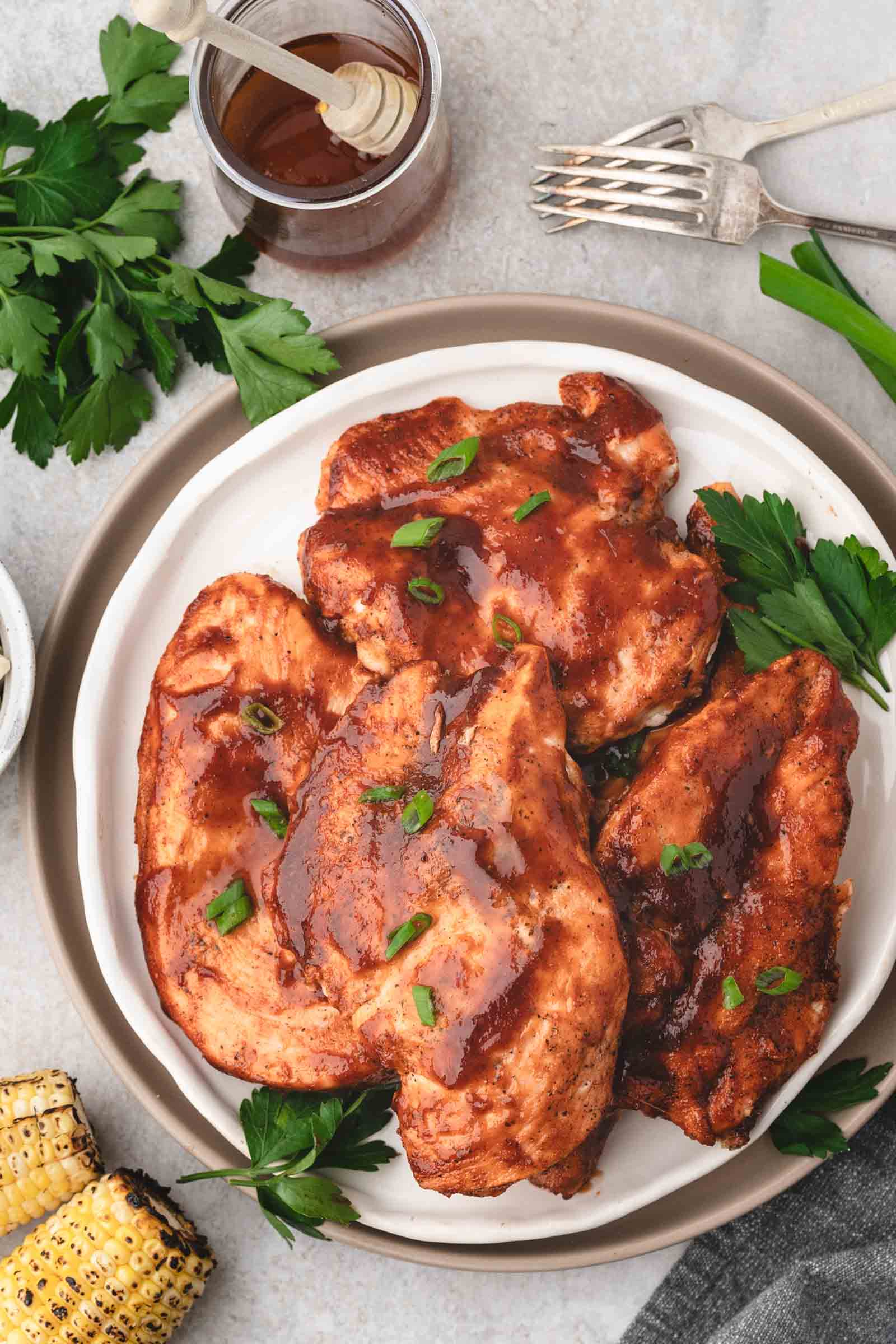 Plate of bbq chicken breasts glazed with barbecue sauce, garnished with parsley and green onions, served with grilled corn and a jar of sauce on the side.
