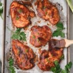Five pieces of cooked barbecue chicken breast on a parchment-lined baking sheet, garnished with chopped herbs, with a jar of sauce and fresh parsley on the side.