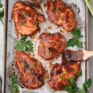 Five pieces of cooked barbecue chicken breast on a parchment-lined baking sheet, garnished with chopped herbs, with a jar of sauce and fresh parsley on the side.