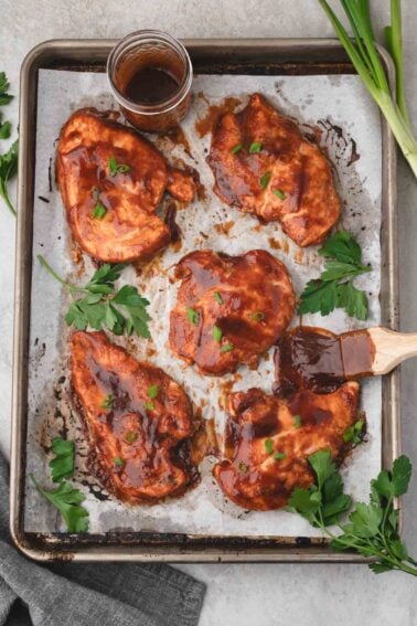 Five pieces of cooked barbecue chicken breast on a parchment-lined baking sheet, garnished with chopped herbs, with a jar of sauce and fresh parsley on the side.