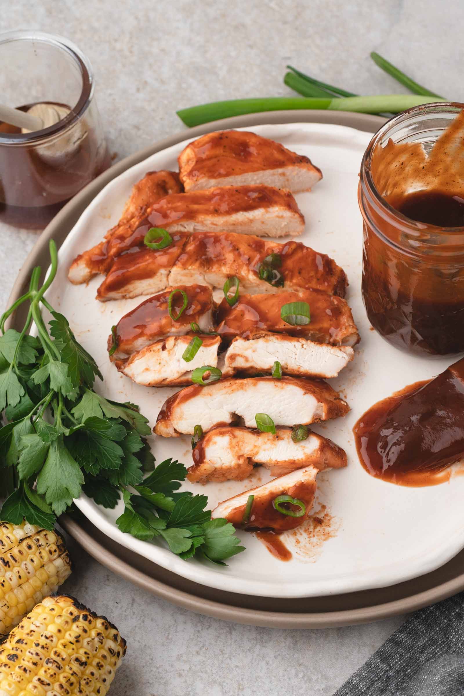 Sliced bbq chicken breast topped with barbecue sauce and green onions on a white plate, served with parsley, corn on the cob, and a jar of extra sauce.