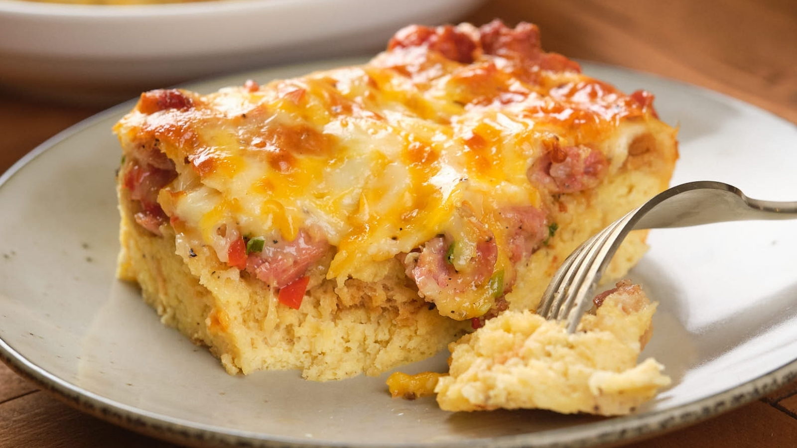 Close-up of a slice of cheesy casserole with bits of meat and vegetables on a plate, with a fork taking a piece.