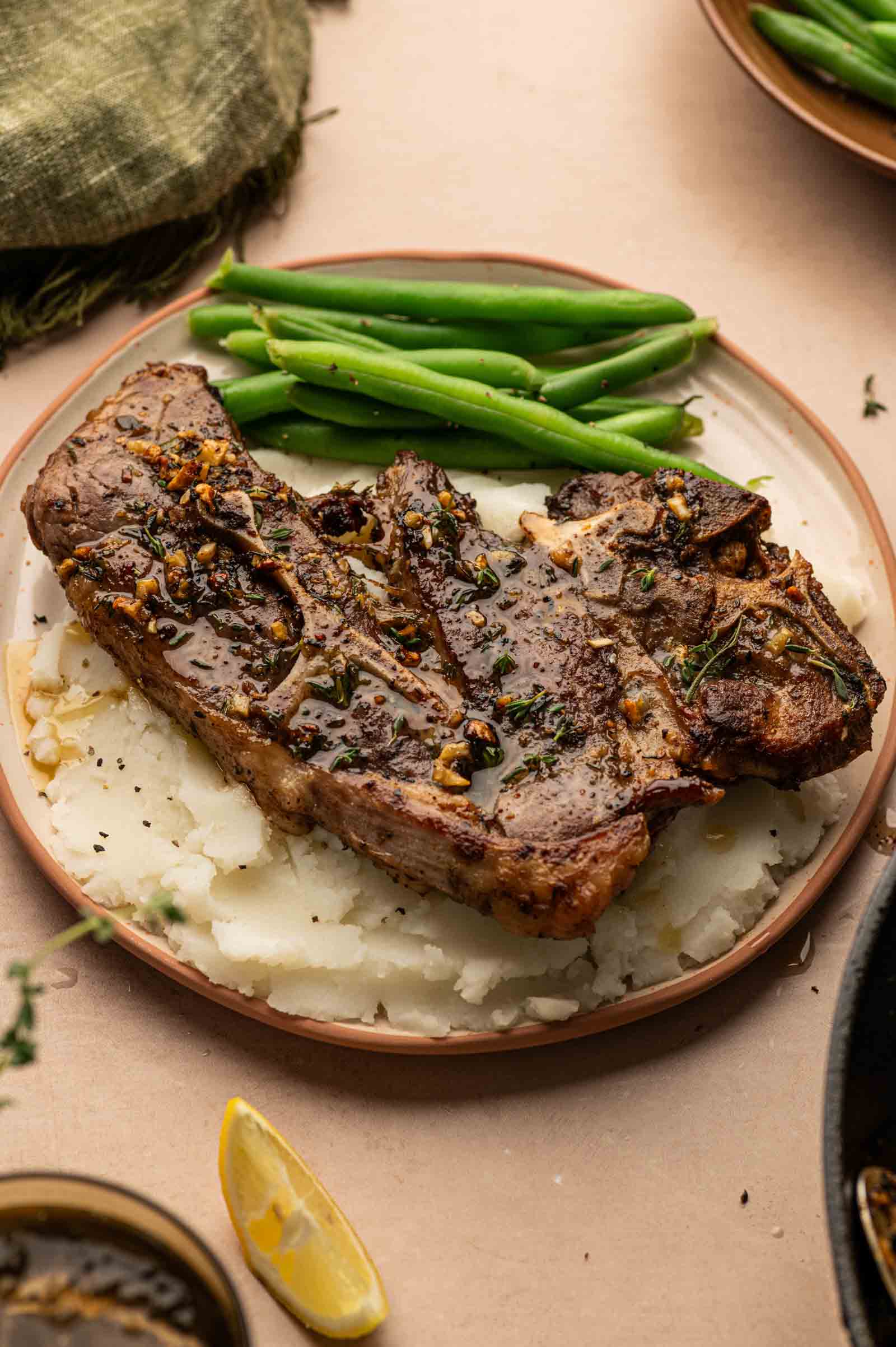 A cooked lamb shoulder chop sits on mashed potatoes, served with green beans on a round plate.