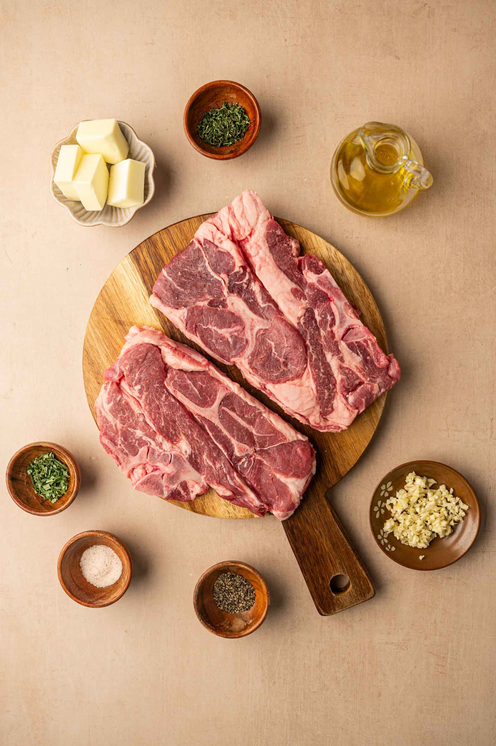 Two raw lamb shoulder chops on a wooden cutting board surrounded by small bowls of butter, oil, chopped herbs, minced garlic, salt, and pepper.